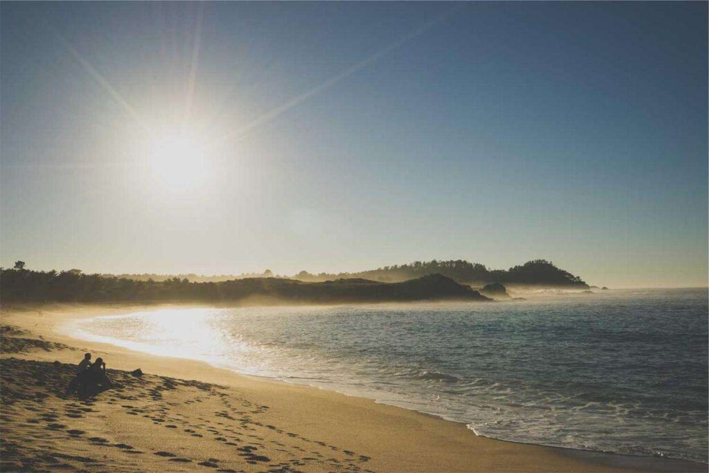A serene beach scene at sunset, with the sun low in the sky casting a warm glow over the sandy shore and gentle waves. A couple sits on the sand, enjoying the view. Forested hills are visible in the background.