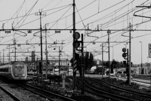 Black and white photo of a train station with multiple tracks and overhead power lines. A train approaches from the left, while signals and poles are visible throughout the scene. Buildings and trees are in the background.