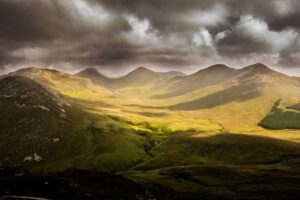 A dramatic landscape featuring rolling green hills and rugged mountains under a cloudy sky. Sunlight breaks through the clouds, casting patches of light on the terrain, creating a striking contrast between light and shadow.