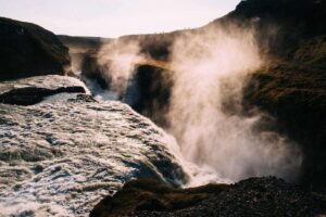 A powerful river cascades through a rocky canyon, creating misty spray as it falls. The landscape is rugged and dramatic, with sunlight illuminating the mist and casting a warm glow over the scene.