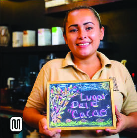 A smiling person holds a colorful chalkboard that reads "Lugar del Cacao" in a cozy café setting. Shelves with artisan cacao and coffee products are visible in the background.