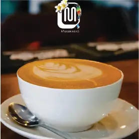 A cup of ethically sourced, organic single origin coffee with latte art sits in a white cup and saucer on a wooden table. A spoon rests on the saucer. The Mishkaao logo with floral design is visible above the cup, highlighting their commitment to quality and ethics.