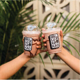 Two hands clink chocolate smoothie cups labeled "MU KA KAO," topped with clear dome lids. In the background of greenery and a patterned wall, enjoy the rich taste of artisan cacao and coffee for a truly delightful experience.