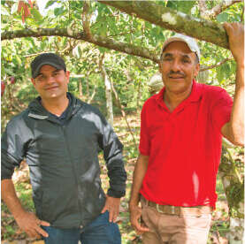 Two men stand in an orchard. The man on the left wears a black jacket and cap, while the man on the right dons a red shirt and white cap. They smile beneath lush greenery, discussing their passion for artisan cacao and coffee cultivated with care for nature's finest offerings.