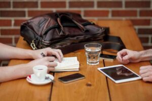 Two people seated at a wooden table with a notebook, smartphone, tablet, and a glass of water. A leather bag rests on the table. The background features a brick wall.