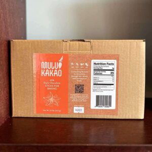 A brown cardboard box labeled "Milk Chocolate Bars" is filled with 65% dark chocolate sticks intended for baking, resting on a wooden shelf. The rich flavor is complemented by a nutrition facts label and a QR code, enhancing the sophisticated appearance of this gourmet box.