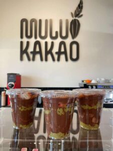 Three plastic cups filled with layered chocolate and green cake sit on a counter. In the background, a sign reads "MULU KAKAO" above coffee machines and cups at this cozy Hattiesburg chocolate & coffee spot.