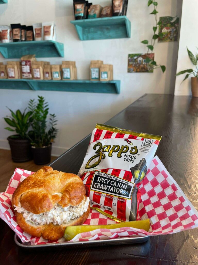 A chicken salad croissant sandwich on red-checkered paper is served with a dill pickle spear and Zapp’s Spicy Cajun Crawtators chips at this charming Hattiesburg cafe, with shelves of packaged goods and green plants in the background.
