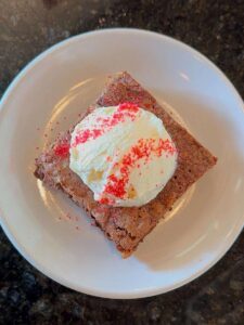 A square brownie from a local coffee and chocolate shop, topped with a scoop of vanilla ice cream and red sprinkles, served on a white plate against a dark countertop background.