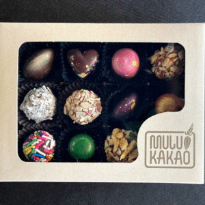 A box of 12 assorted artisan bonbons from the Mulukakao Bonbon Collection features chocolates and truffles in unique shapes and colors, in a light box with a clear window and "MULU KAKAO" branding at the bottom right corner.