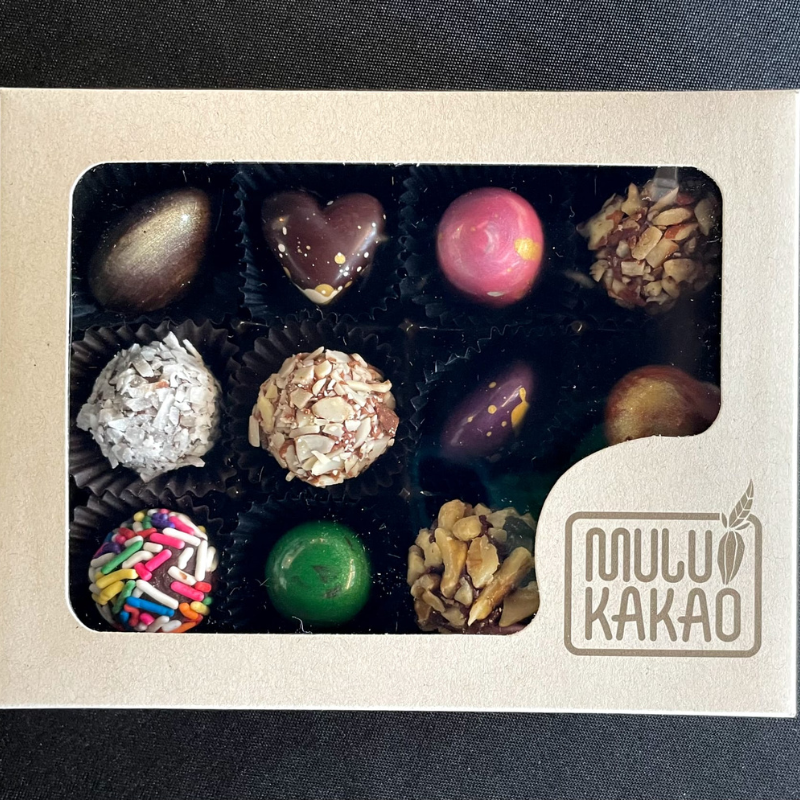 A box of 12 assorted artisan bonbons from the Mulukakao Bonbon Collection features chocolates and truffles in unique shapes and colors, in a light box with a clear window and "MULU KAKAO" branding at the bottom right corner.