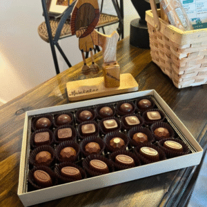 The Mulukakao Signature Boozy Bonbon Collection – 24-Piece Gift Edition is displayed on a wooden table beside a small wooden figurine and wicker basket, with each bonbon neatly arranged in its own wrapper.