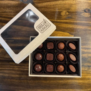 An open Mulukakao Signature Boozy Bonbon Collection – 24-Piece Gift Edition sits on a wooden table, revealing rows of round, square, and oval chocolates in black cups. The box’s clear window lid rests nearby.