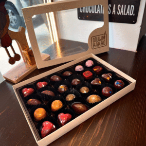 A Box of 12 Bonbons from the Mulukakao Bonbon Collection, in individual cups, sits open on a dark wooden table near a window. The lid with "Mulu Kakoa" branding and clear window rests behind the artisan chocolates.